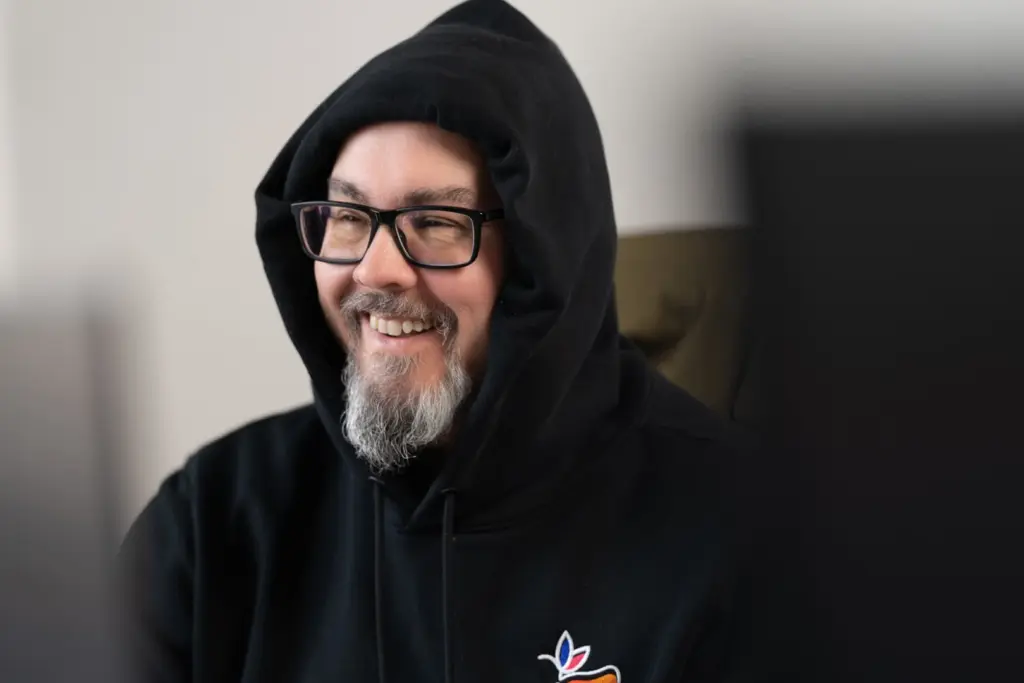 Mann mit schwarzer Kapuzenjacke und Brille, lächelnd, mit grauem Bart vor hellem Hintergrund.