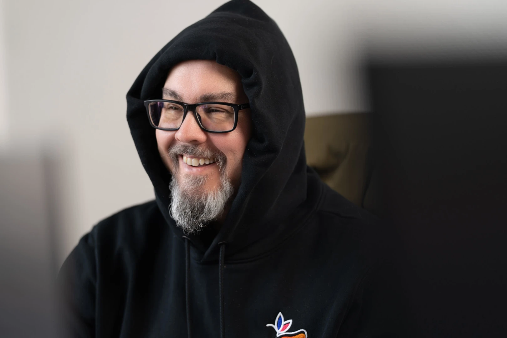 Kontakt Mann mit schwarzer Kapuzenjacke und Brille, lächelnd, mit grauem Bart vor hellem Hintergrund.