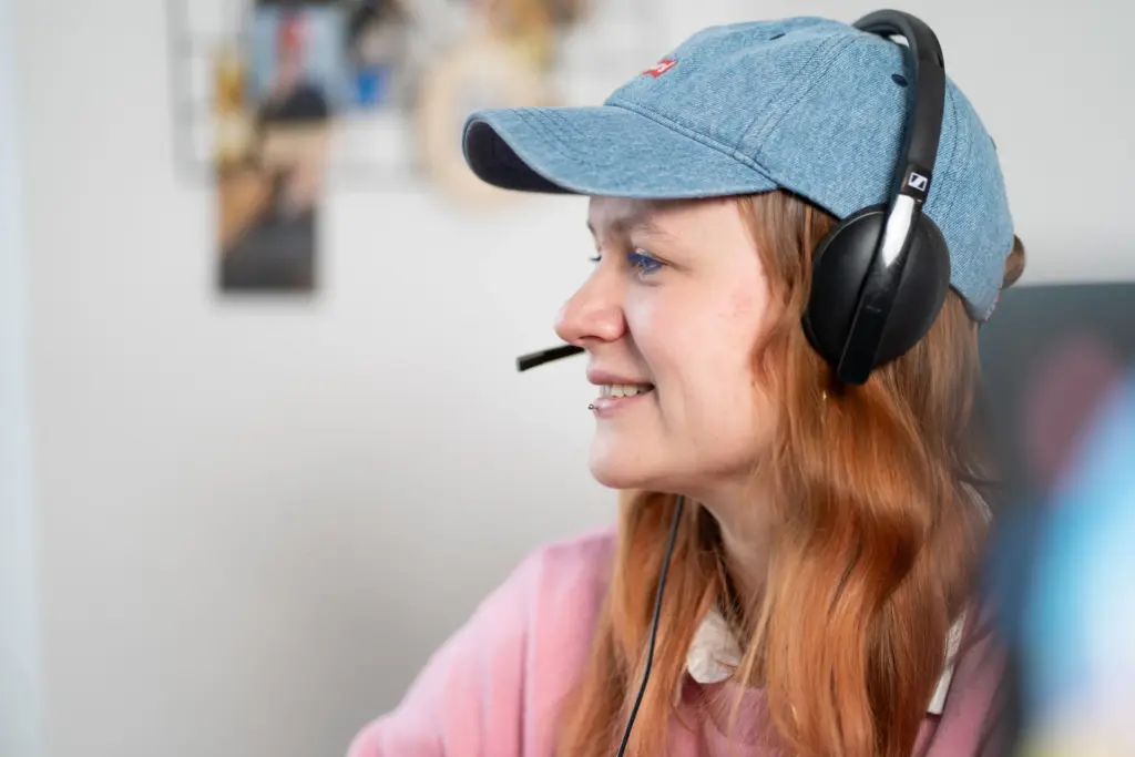 Frau mit roten Haaren, blauer Kappe und Kopfhörer mit Mikrofon, lächelnd seitlich nach rechts blickend.