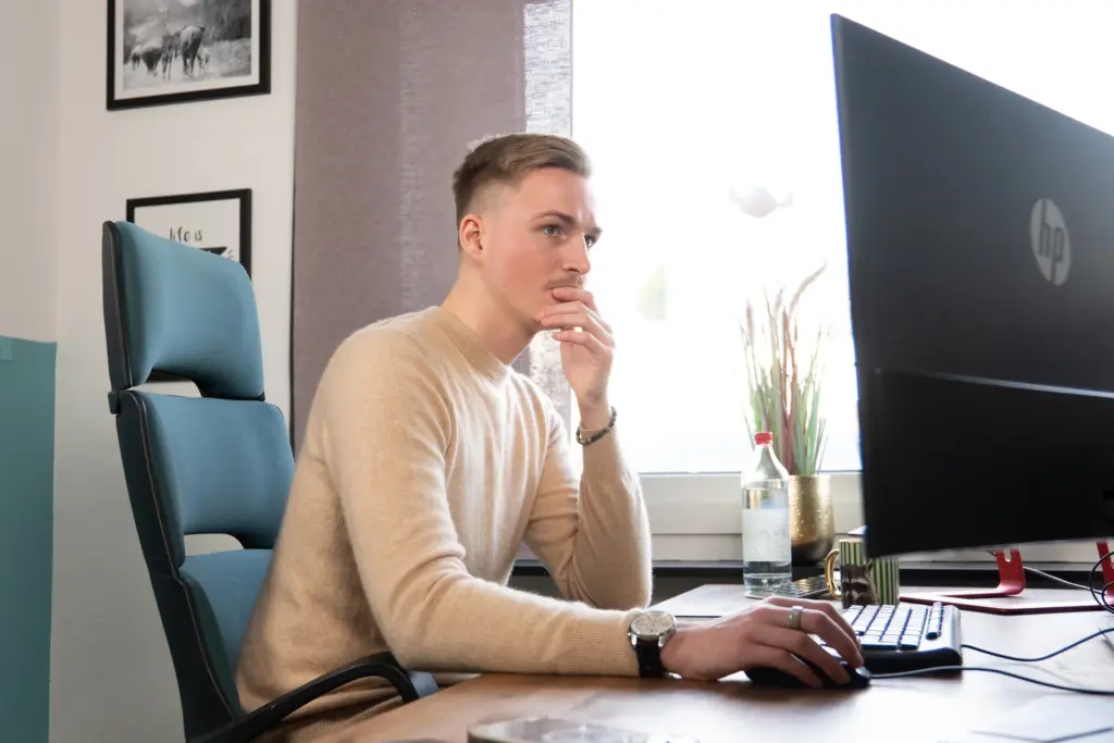 Junger Mann in beige Pullover sitzt nachdenklich vor Computerbildschirm an einem Schreibtisch mit Wasserflasche und Kaffeetasse.