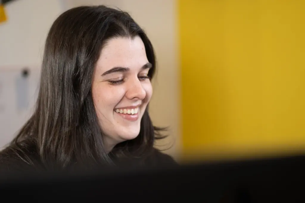 Frau mit dunklen langen Haaren lächelt, vor gelbem Hintergrund, Blick leicht nach unten gerichtet.