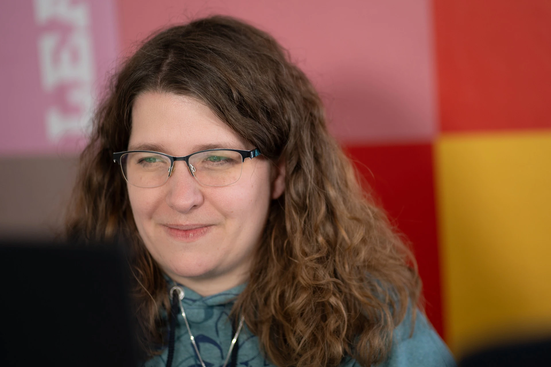 Kontakt Frau mit langen lockigen braunen Haaren und Brille blickt lächelnd auf einen Bildschirm vor ihr, bunte Wand im Hintergrund.
