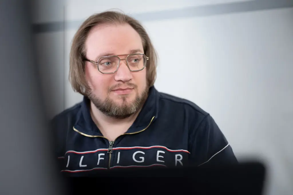 Mann mit Brille und Bart schaut nach rechts, trägt dunkelblauen Pullover mit Reißverschluss und Aufschrift "TOMMY HILFIGER".