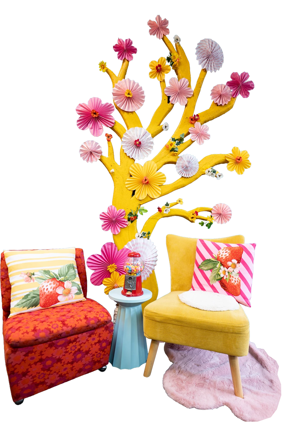 Gelber Stuhl mit rosa Erdbeerkissen neben rotem Stuhl mit gestreiftem Erdbeerkissen, kleinem blauen Tisch mit Kaugummiautomat und gelb bemaltem Baum mit Papierblumen dekoriert.