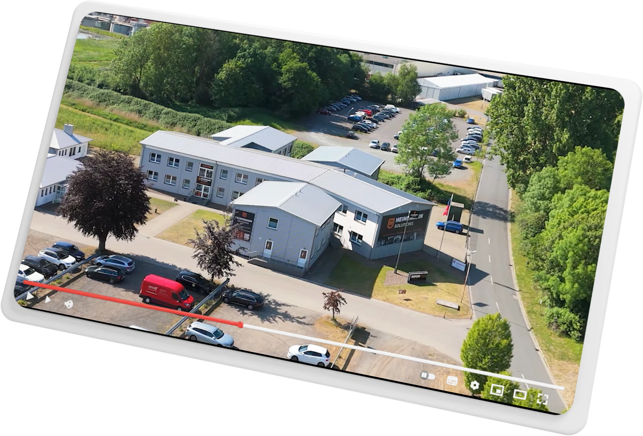 Luftaufnahme eines Bürogebäudes mit Parkplatz, umgeben von Bäumen und Straßen, mit mehreren geparkten Autos.